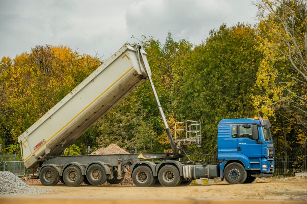 A blue dump truck unloading at a construction site surrounded by autumn foliage.